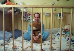 The brown teddy bear is being held by a man who is seated in the crib.