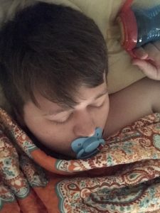 She is sleeping in bed with a baby man who is sucking the baby's nipple.