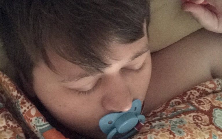 She is sleeping in bed with a baby man who is sucking the baby's nipple.