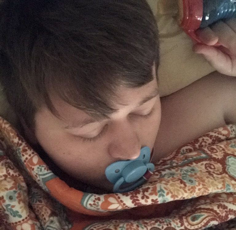 She is sleeping in bed with a baby man who is sucking the baby's nipple.