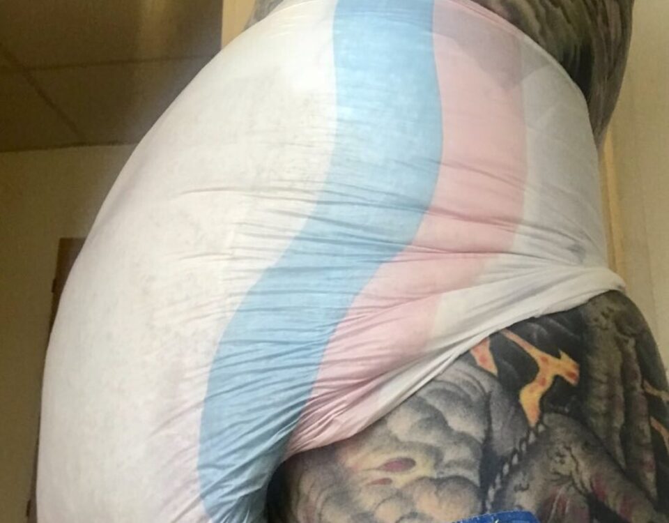 A handsome man is showing her sexy diaper while wearing the diaper and sporting a tattoo. For the photo session, he is standing.